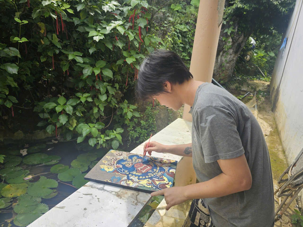 Định Tâm đứng vẽ bức thangka Đại Hắc Thiên trên bệ đá bên hồ sen, cây lá rậm phía sau